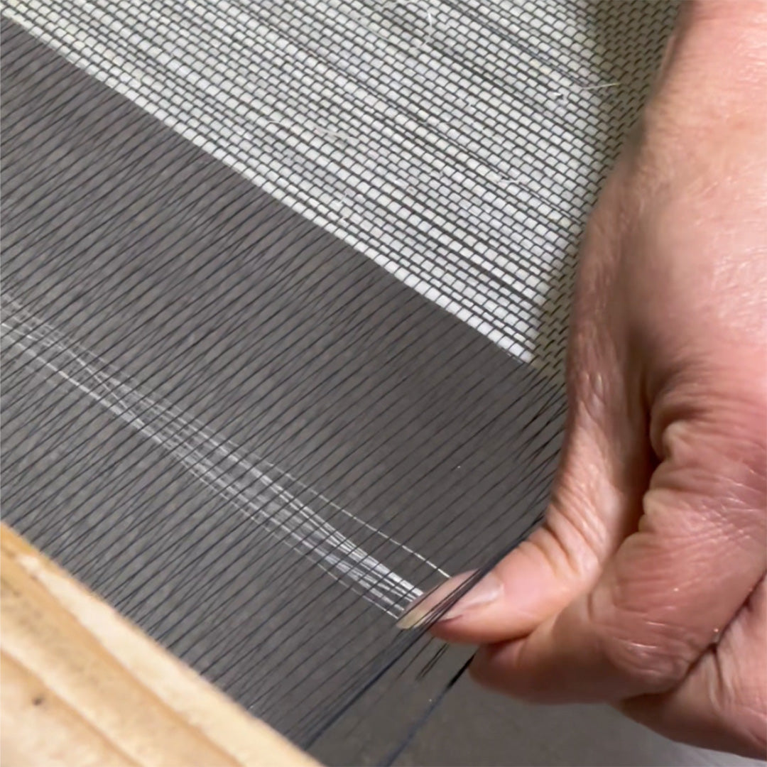 Close-up of a hand weaving grasscloth fibers on a loom, creating a detailed textile pattern with a contrasting white woven section in the background.