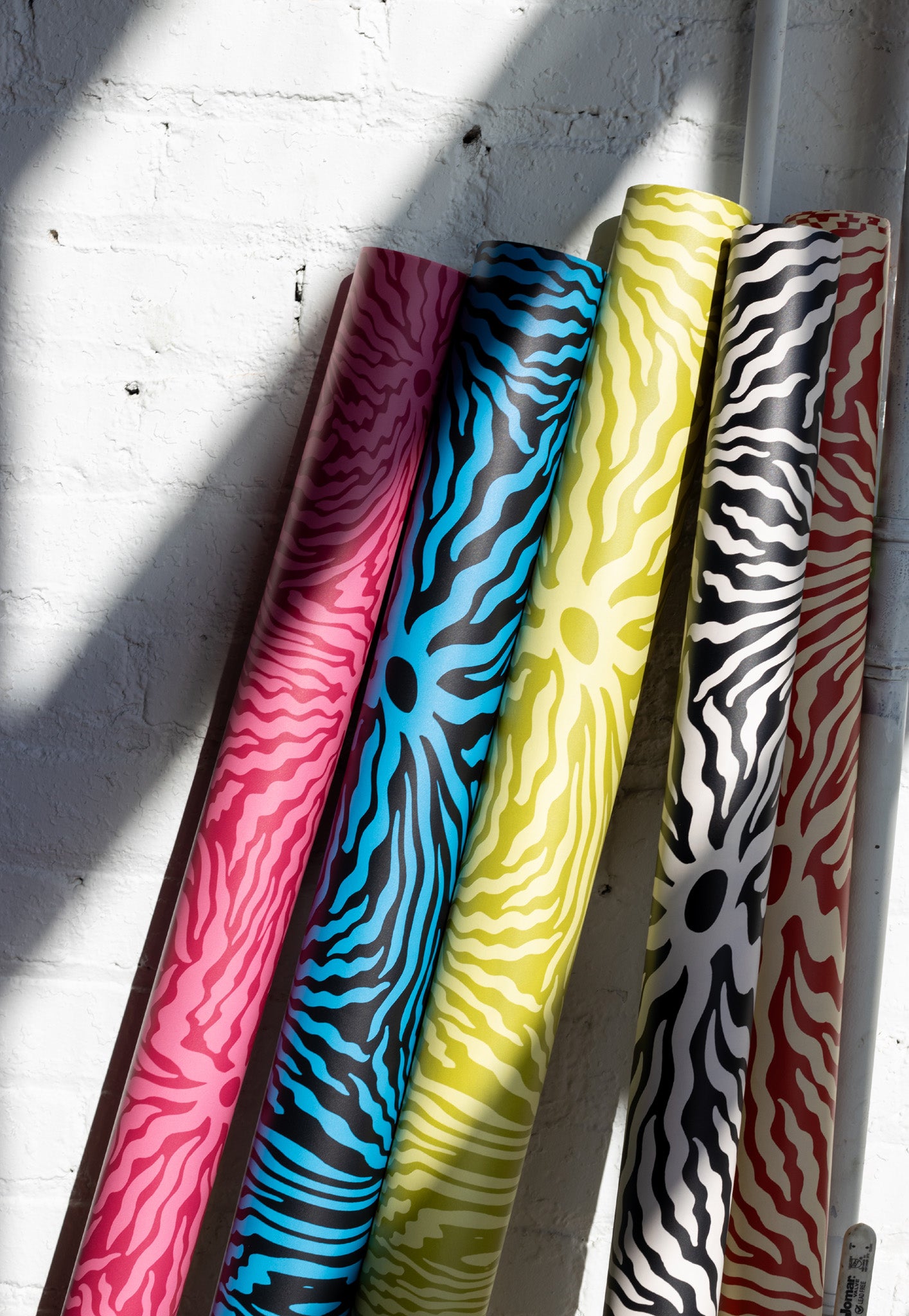 Close-up of colorful patterned wallpaper rolls in pink, blue, yellow, and black,
leaning against a white brick wall with natural light casting shadows.