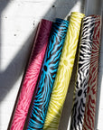 Close-up of colorful patterned wallpaper rolls in pink, blue, yellow, and black,
leaning against a white brick wall with natural light casting shadows.