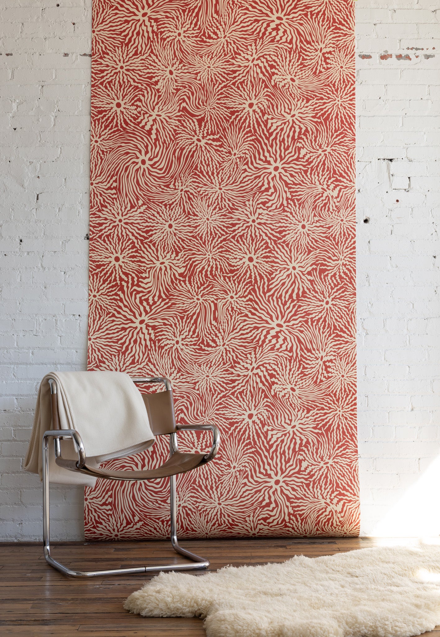 Stylish tan leather and chrome chair draped with a white blanket, placed in front of
red patterned wallpaper with abstract swirling design, on a fluffy cream rug.