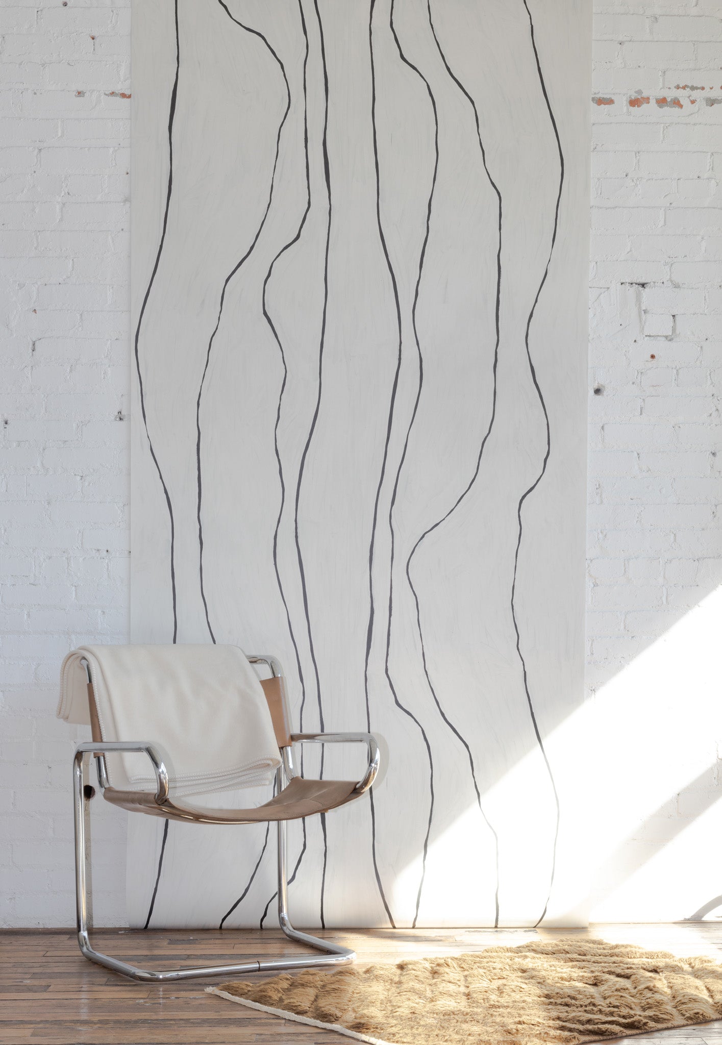 Off-white wallpaper with black hand-painted vertical lines, paired with a chrome and leather sling chair and a textured beige rug.