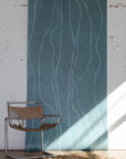 Dark teal wallpaper with subtle wavy vertical lines in light blue, styled with a modern leather and chrome chair on a textured beige rug.