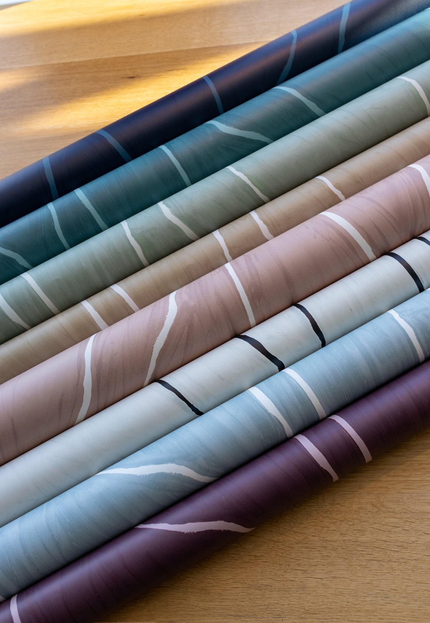 Rolled wallpaper samples in various colors, including navy, teal, green, beige, blush, and plum, all featuring organic vertical line patterns, placed on a light wood surface.